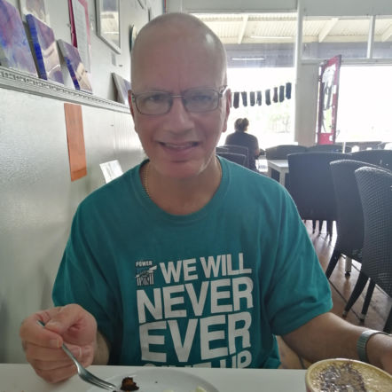 Craig is wearing a teal Port Adelaide shirt and sitting down at a table having just finished some food. He has a fork in his right hand and is smiling at the camera.