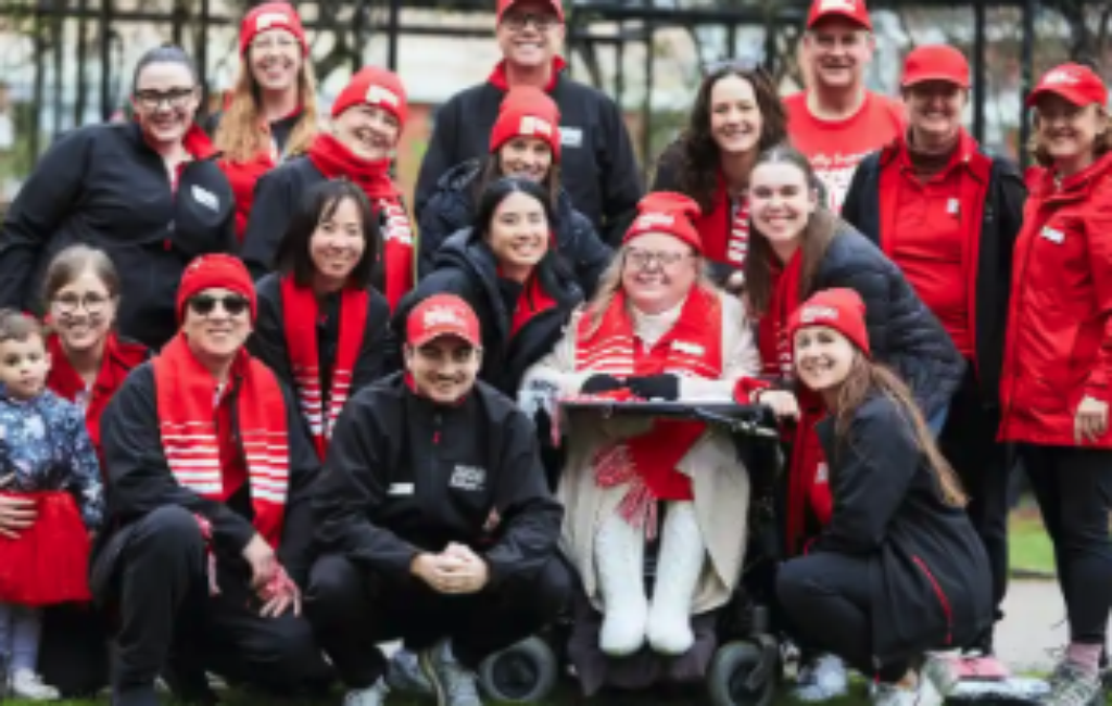A group of people with red beanies and scarves are all smiling at the camera some of them are in wheelchairs