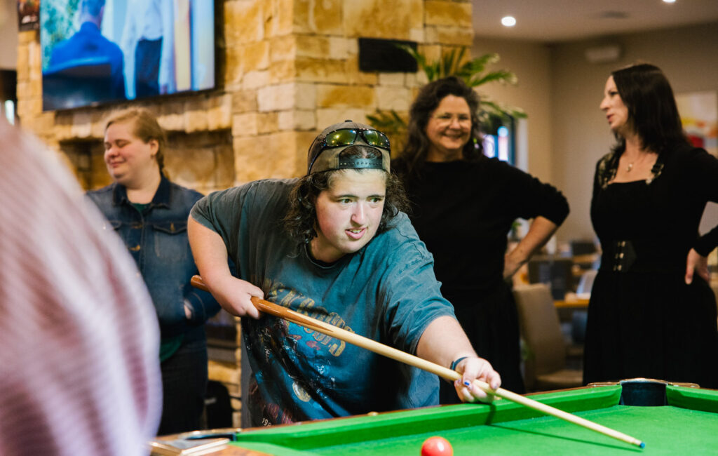A person playing pool with a group of people socialising in the background at a bar. The people are smiling and happy