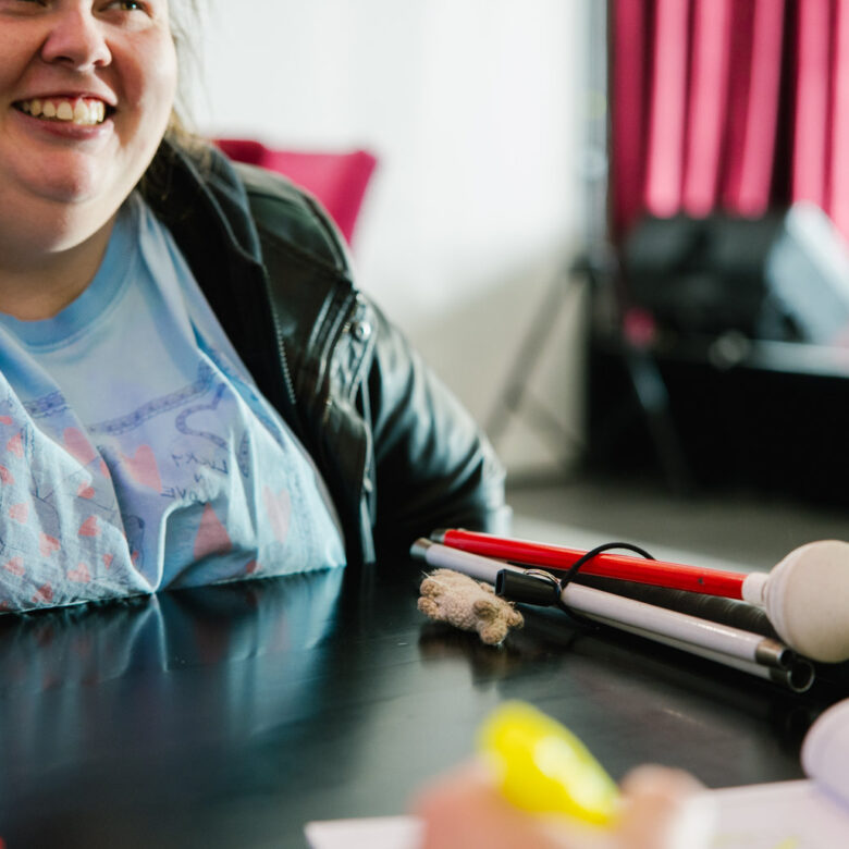 A woman is sitting talking to someone unseen across a table. she has a cane folded up on the table in front of her.