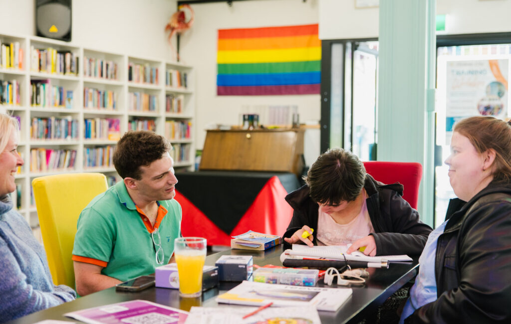 A group of people are at a table doing activities. There is a rainbow pride flag on the wall behind them.