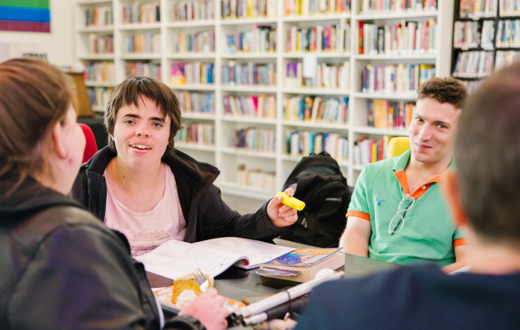people sitting around a table. there is a bookshelf in the background and a woman is smiling at the camera
